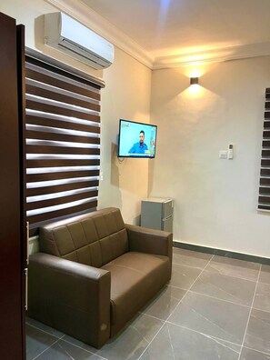 A room with a brown leather sofa, a mounted TV, an air conditioning unit, and a small cabinet. - Newly renovated house in a gated community (ACCRA)