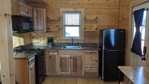 A wooden cabin-style kitchen with a stove, sink, refrigerator, and a window above the sink.