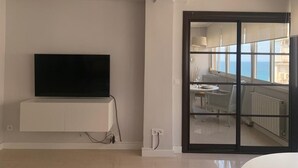 Een moderne woonkamer met een flatscreen-televisie bevestigd aan een wit kastje, een schuifdeur die leidt naar een balkon met uitzicht op zee.