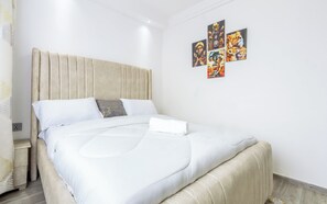 Cama bem arrumada com cabeceira bege, roupa de cama branca e uma pequena mesa de cabeceira.