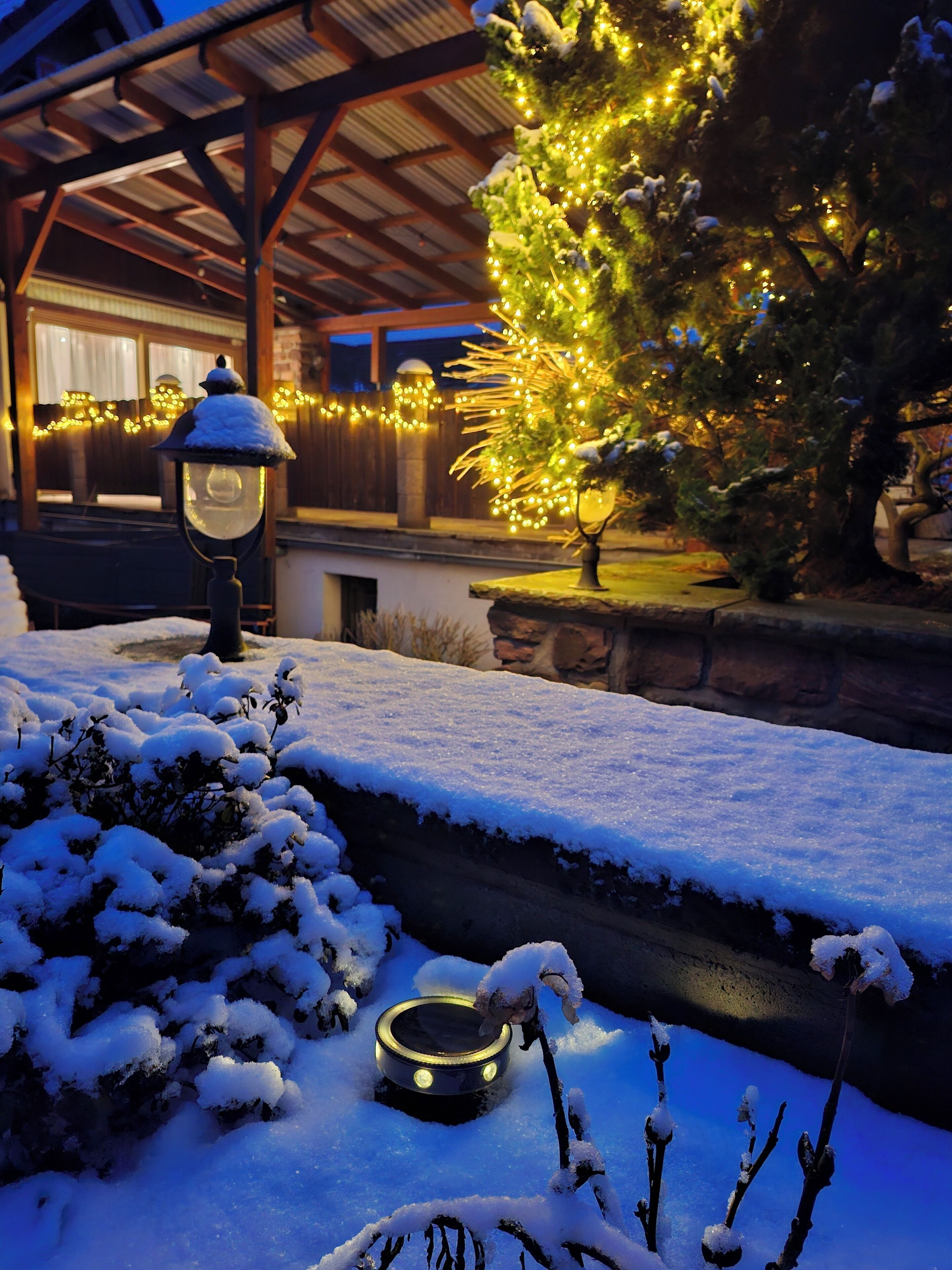 Ein mit Schnee bedeckter Garten mit Laterne, Weihnachtsbaum und Steinmauer.