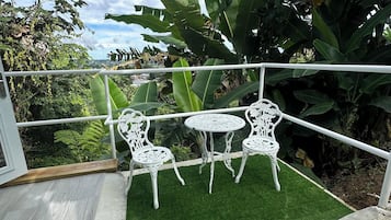 A balcony with white metal chairs and a table, surrounded by lush green plants and a view of the city.