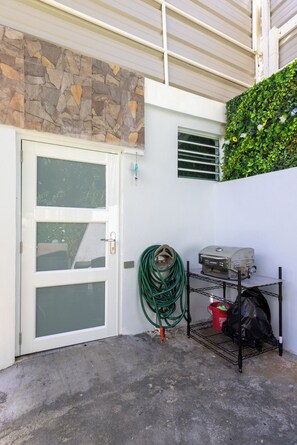 Un garage avec un mur en pierre, un mur végétal, une porte blanche et une étagère en métal sur laquelle se trouvent un gril et un tuyau d’arrosage.