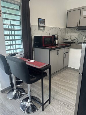 Cocina compacta con microondas, cafetera y un refrigerador pequeño. Hay dos sillas con un mantel individual rojo, una ventana con persianas y un cuadro en la pared.