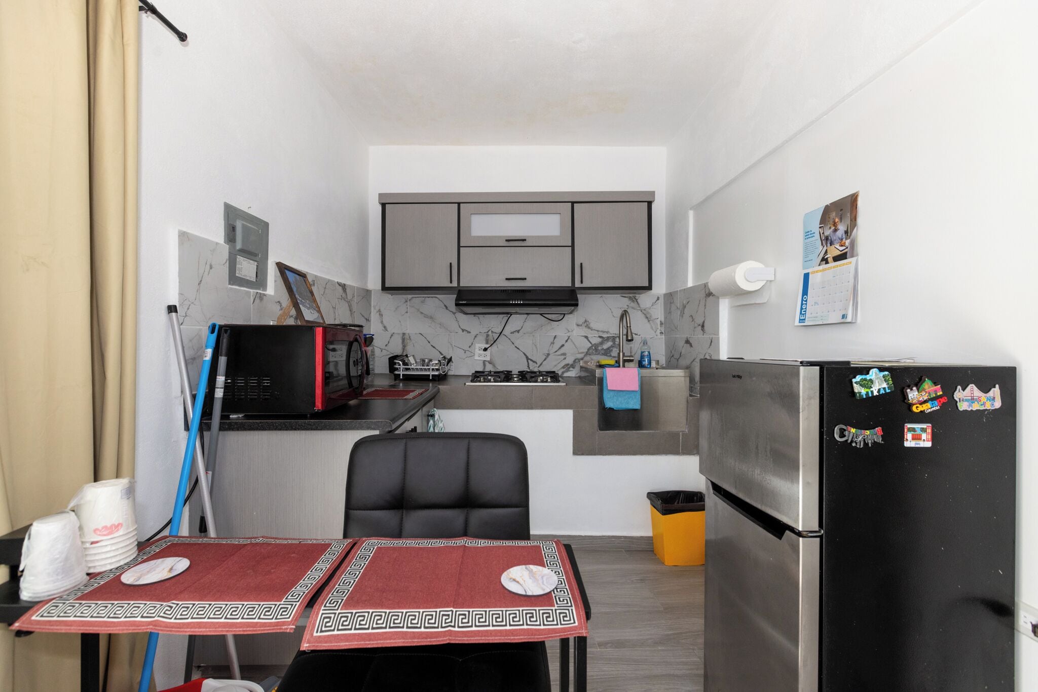 A compact kitchen with grey cabinets, a black refrigerator, and a dining area with a red tablecloth.