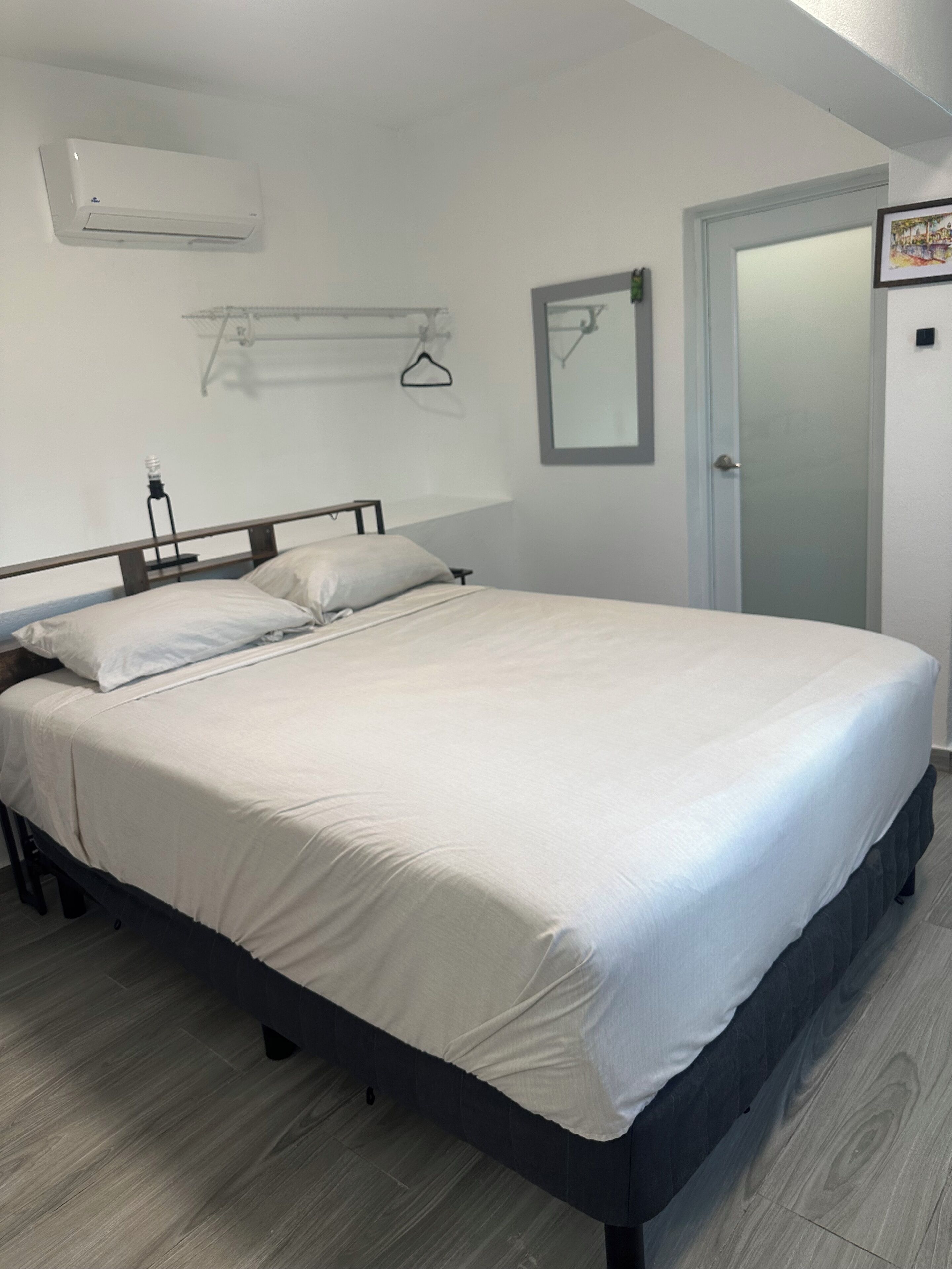 A neatly made bed in a room with a wooden floor, a white air conditioner, a coat rack, and a door with a frosted glass panel.