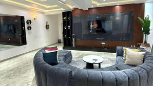 A modern living room with a curved grey sofa, a flat-screen TV mounted on the wall, and a small coffee table. - Kair Serviced Apartment (Benin City)