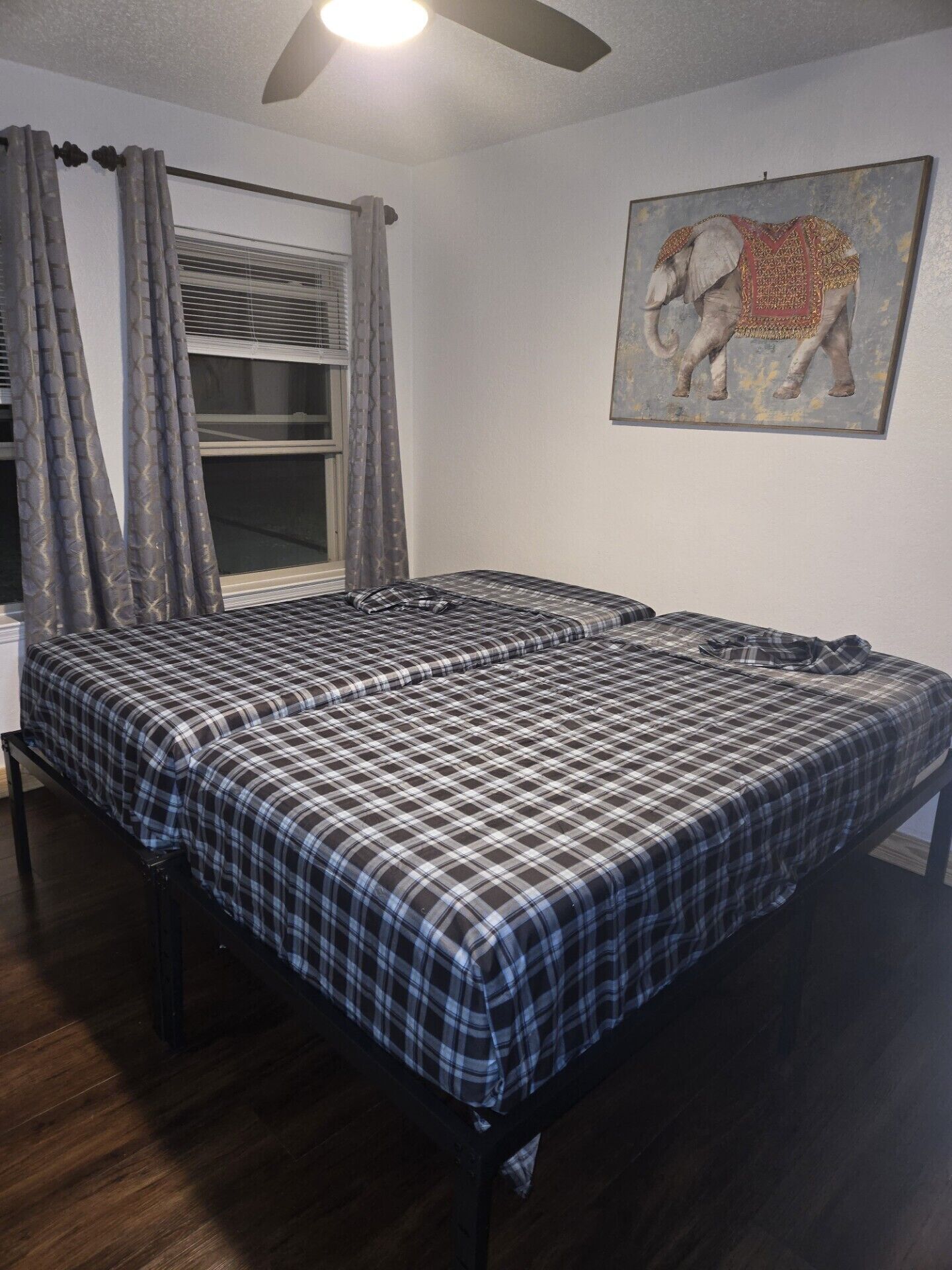 A bedroom with a large bed, a ceiling fan, a window with blinds, and a painting of an elephant on the wall.