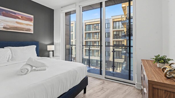 Ein modernes Schlafzimmer mit einem großen Bett, einem Nachttisch, einem Fenster mit Blick auf ein Gebäude und einer hölzernen Kommode.