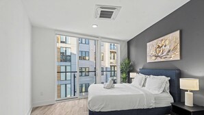 Ein Schlafzimmer mit Bett, Nachttisch, Lampe und einem großen Fenster mit Blick auf Gebäude.