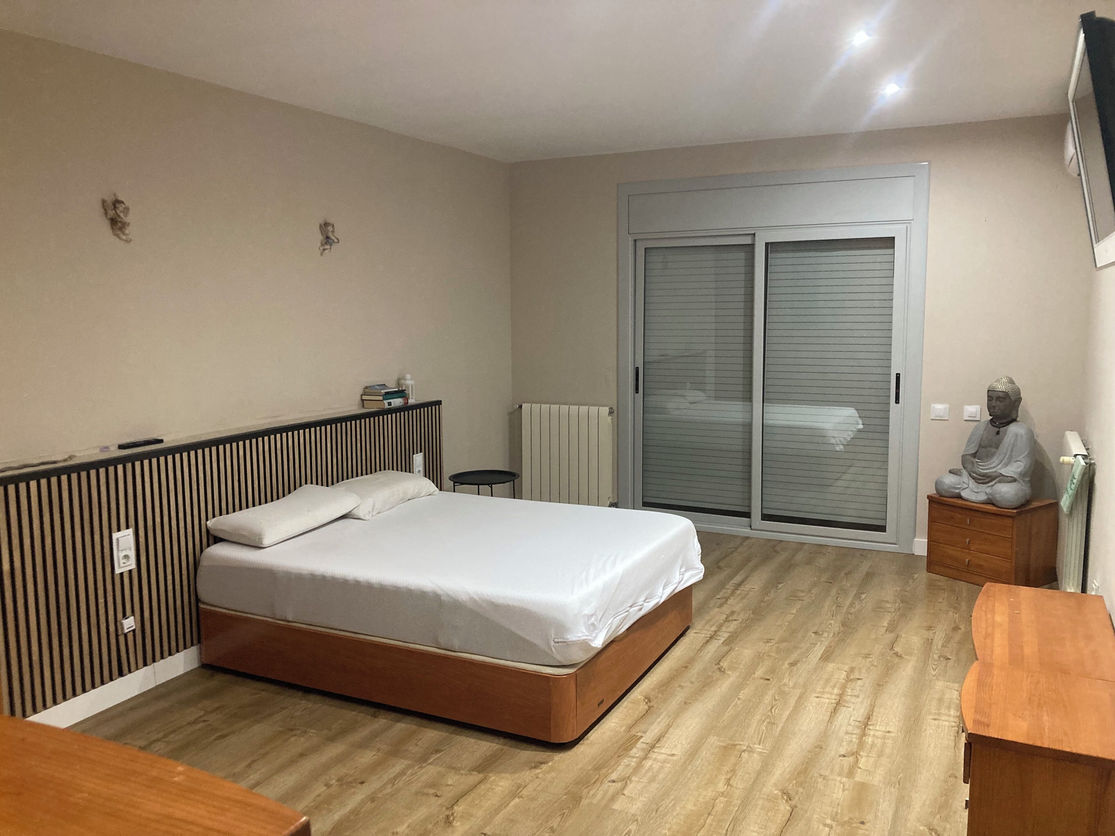 Een slaapkamer met een bed, een houten dressoir, een beeld en een schuifdeur met glas.