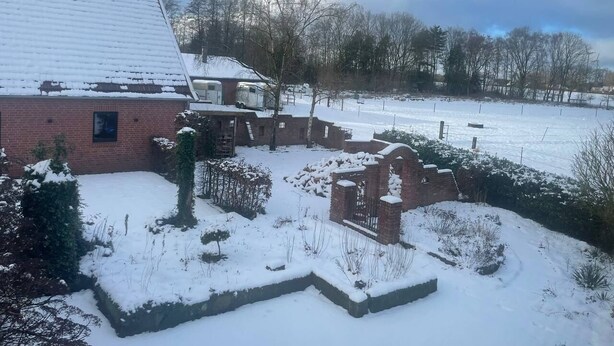 Área externa coberta de neve, com um edifício de tijolos, um portão e um quintal cercado.