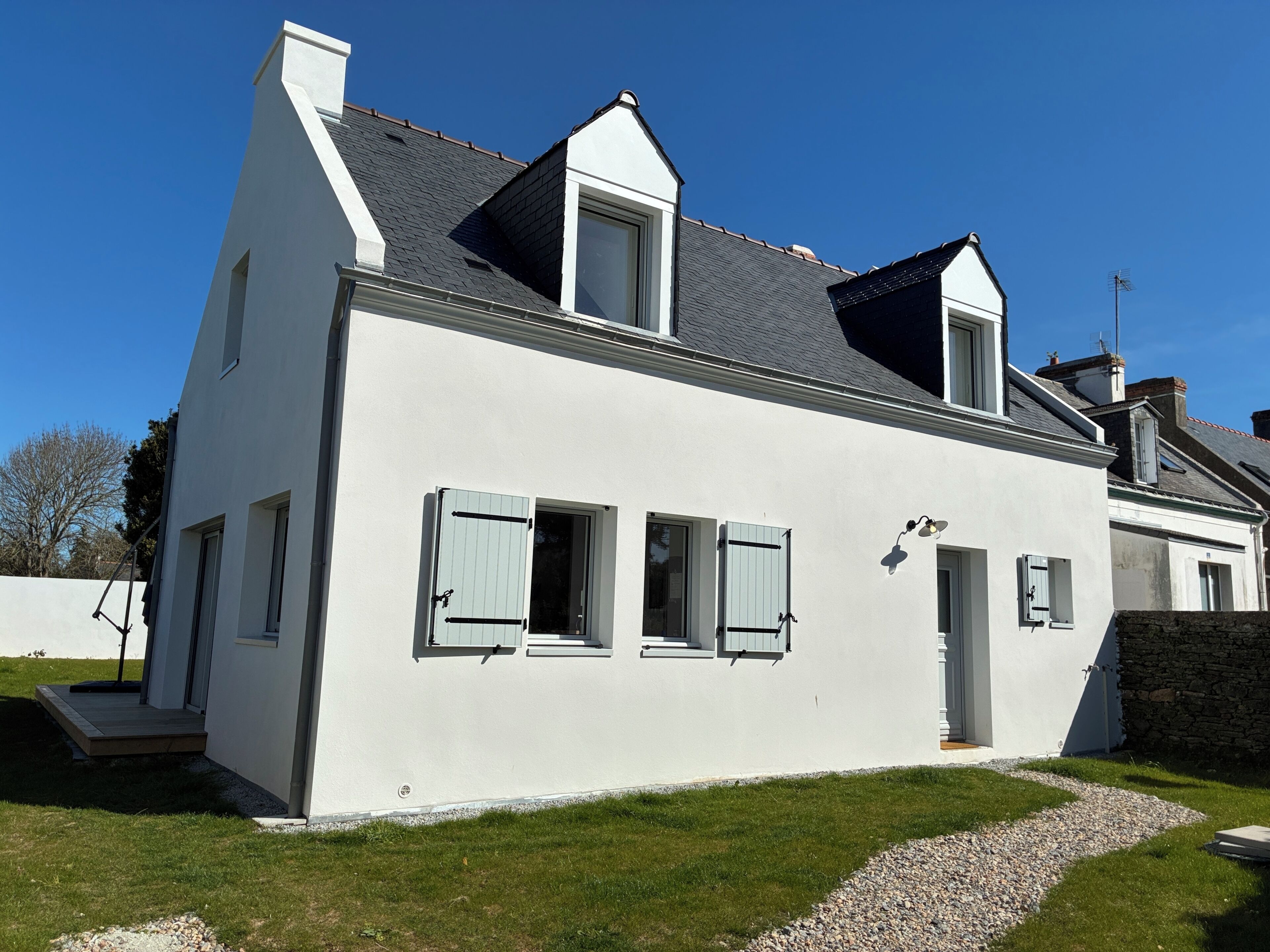 A white building with a sloped roof, multiple windows with shutters, and a gravel pathway leading to the entrance.