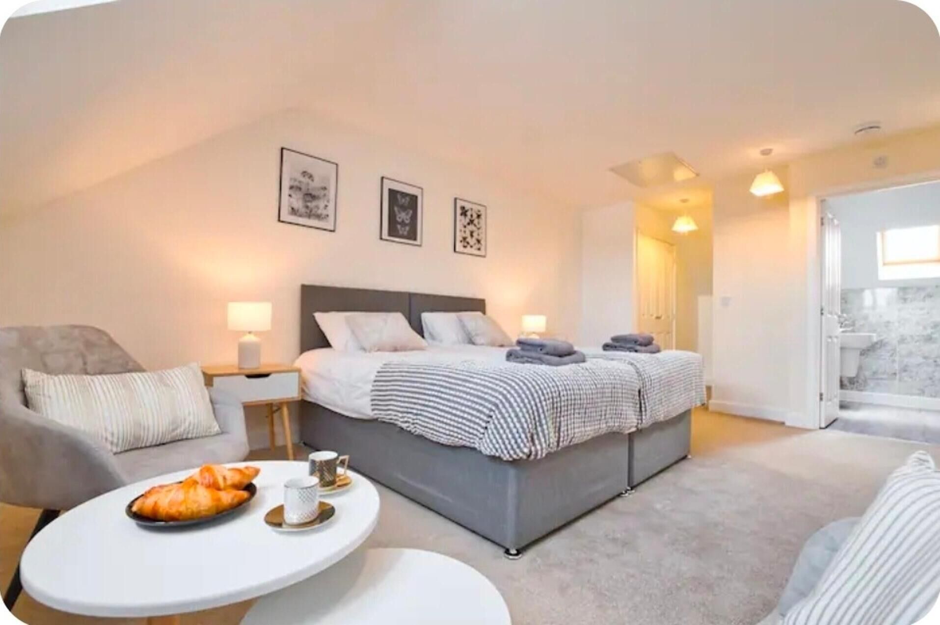 A modern bedroom with a large bed, a sofa, a round table with a plate of croissants, and a bathroom visible through an open door.