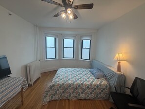 A bedroom with a bed, a desk, a chair, a ceiling fan, and three windows.