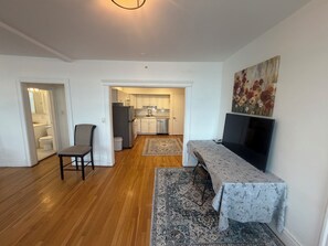 A living room with a TV, a dining table, a chair, and a kitchen in the background.