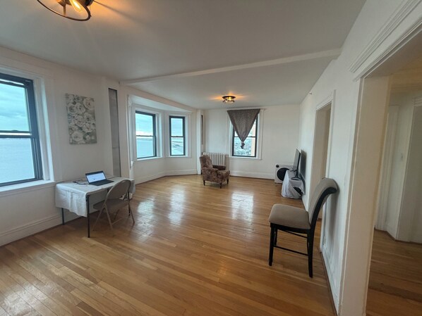 A spacious room with hardwood flooring, a desk with a laptop, a chair, a radiator, and a television.