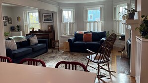 A living room with a dark blue sofa, a wooden rocking chair, and a dining table with red chairs. There are windows with shutters, a piano, and various decorations on the walls. - Spacious 5-bedroom home with private yards - near Brown/RISD (Providence)
