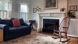 A living room with a dark blue sofa, a wooden rocking chair, a fireplace, and a large window with shutters. - Spacious 5-bedroom home with private yards - near Brown/RISD (Providence)