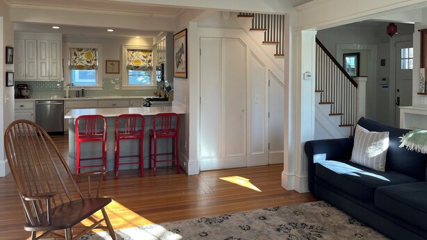 A spacious living area with a dark-colored sofa, a wooden chair, and a kitchen area with white cabinets and red bar stools. - Spacious 5-bedroom home with private yards - near Brown/RISD (Providence)
