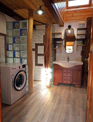 A laundry room with a washing machine, wooden flooring, a tiled wall with a geometric pattern, a wooden ceiling, and a mirror above the sink.