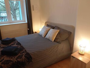 Una cama con un edredón estampado, almohadas, una lámpara de noche y una ventana con vistas a edificios y árboles.