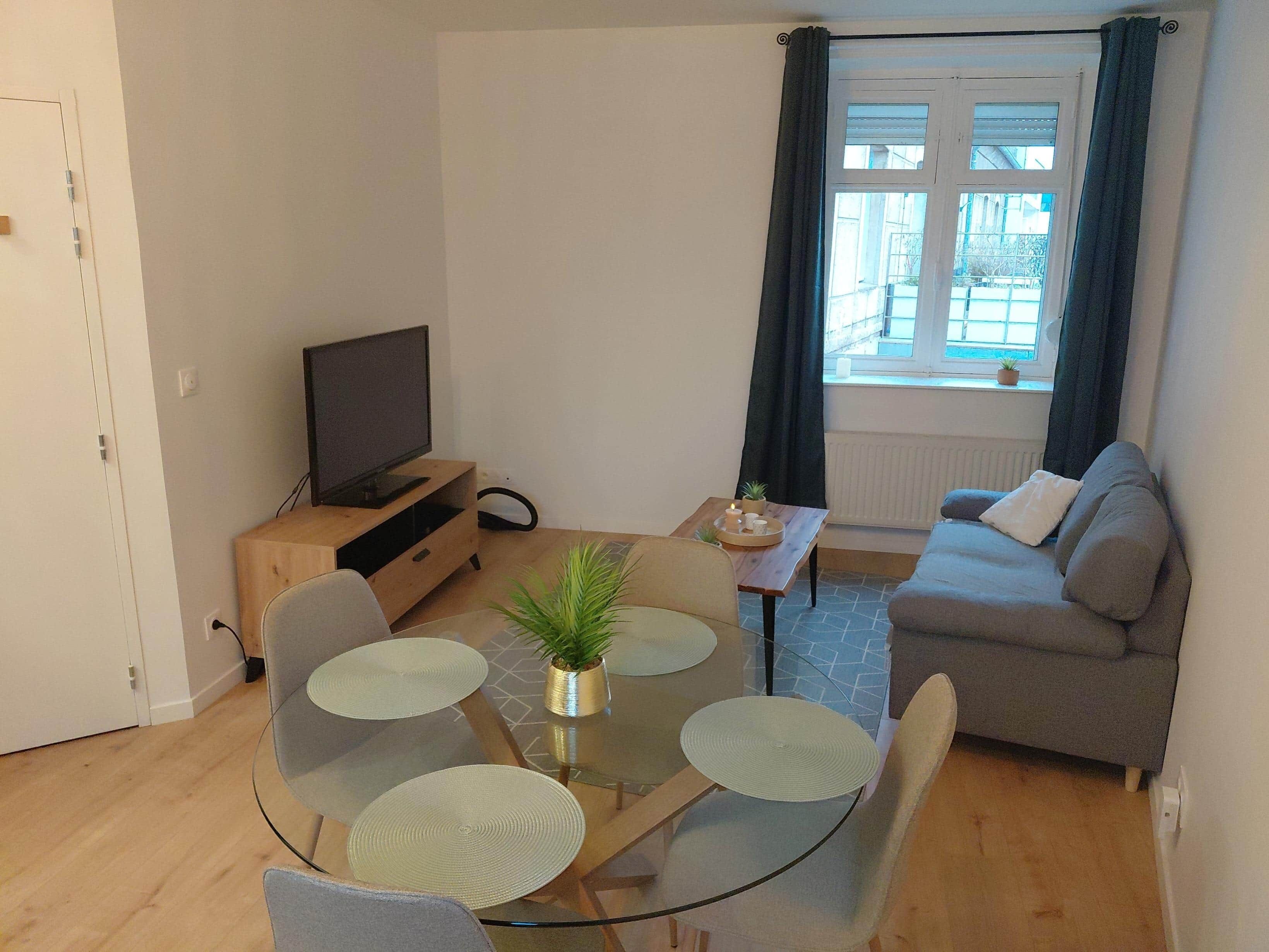 Una sala de estar con sofá, televisión, mesa de vidrio con sillas y ventana con cortinas.