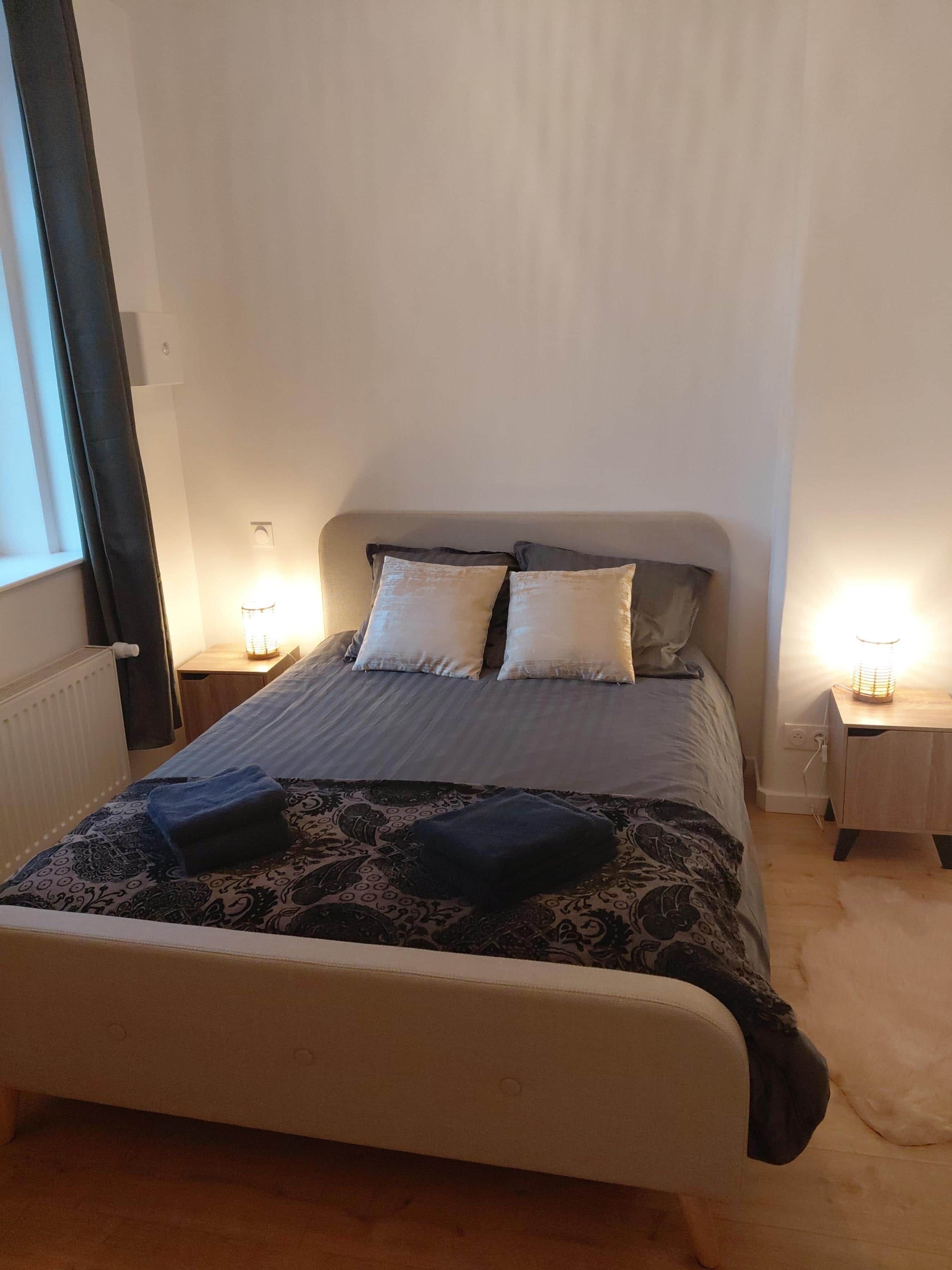 Un lit bien fait, recouvert d’une couvre-lit à motifs et agrémenté de deux oreillers, flanqué de deux tables de chevet avec des lampes, dans une pièce aux murs unis et dotée d’une fenêtre avec des rideaux.