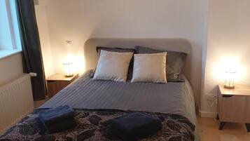 Un lit bien fait, recouvert d’une couvre-lit à motifs et agrémenté de deux oreillers, flanqué de deux tables de chevet avec des lampes, dans une pièce aux murs unis et dotée d’une fenêtre avec des rideaux.
