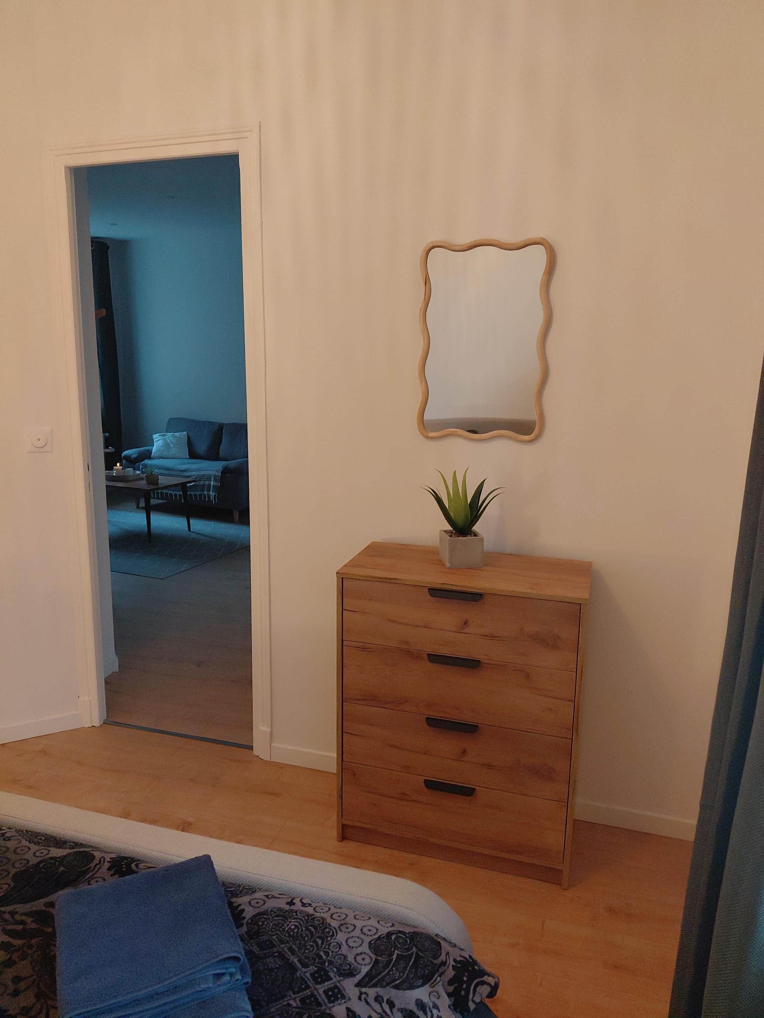 Un dormitorio con cama, cómoda de madera con cajones, planta pequeña en maceta y espejo en la pared.