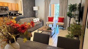 Una sala moderna con un comedor de madera oscura, un sofá y dos sillones. Se ve una cocina con armarios de madera y un microondas al fondo.