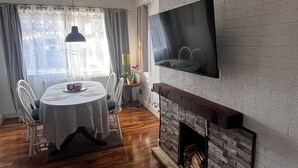 Ein Wohnzimmer mit Kamin, Wandfernseher, Esstisch mit Stühlen und einem fenster mit Vorhängen.