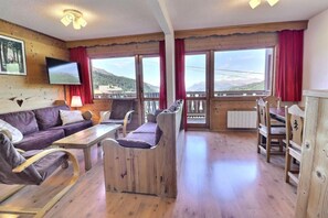 A room with a wooden floor, a sofa, a table, chairs, a TV mounted on the wall, and large windows with a view of mountains. - 150m from ski slopes, 3rd floor, balcony, 53m², Méribel-Mottaret (Méribel - Les Allues)
