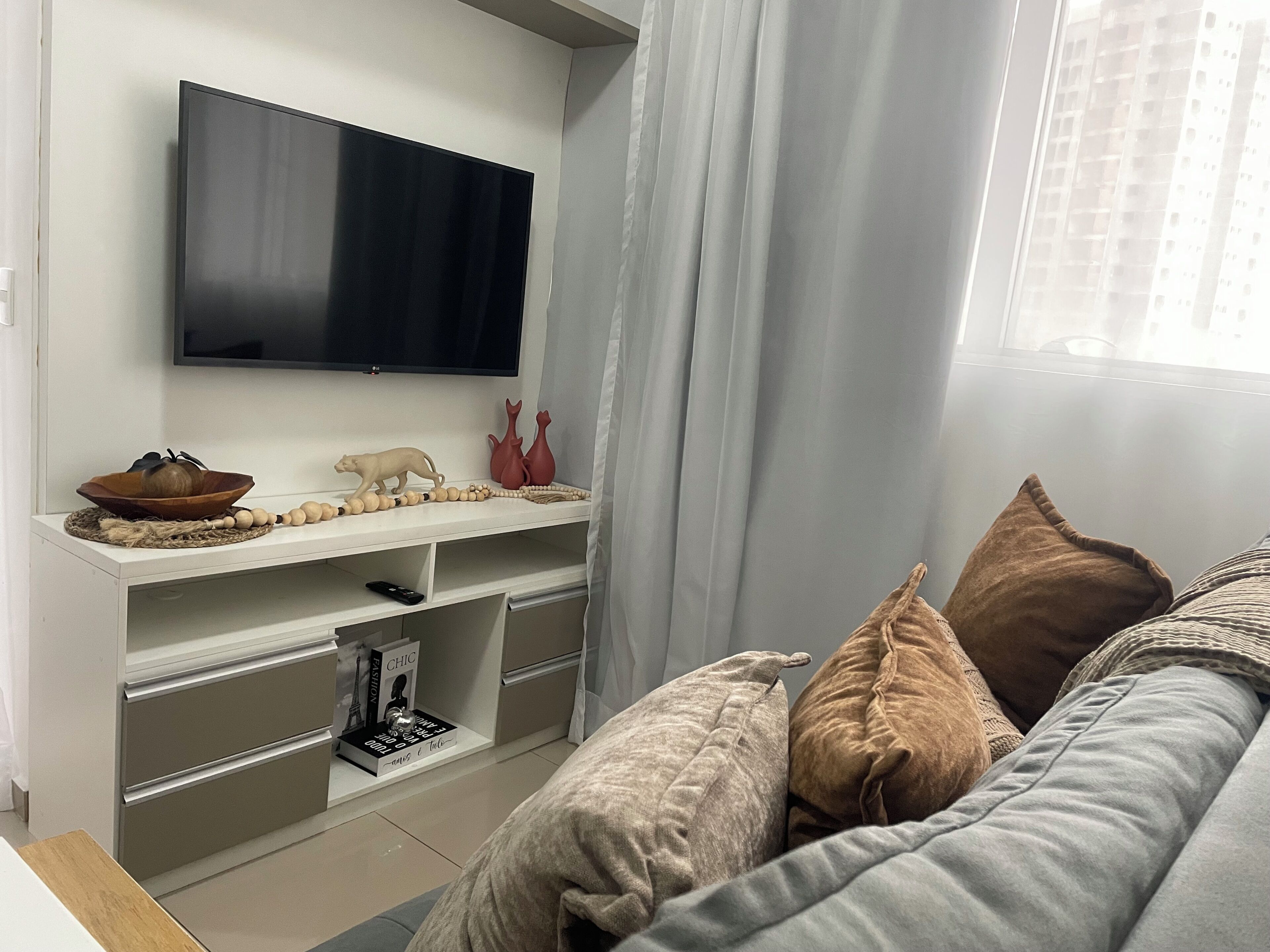 Uma sala de estar moderna com uma TV de tela plana, uma estante de madeira com objetos decorativos, um sofá com almofadas e uma janela com cortinas.