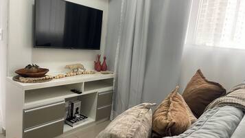 Uma sala de estar moderna com uma TV de tela plana, uma estante de madeira com objetos decorativos, um sofá com almofadas e uma janela com cortinas.