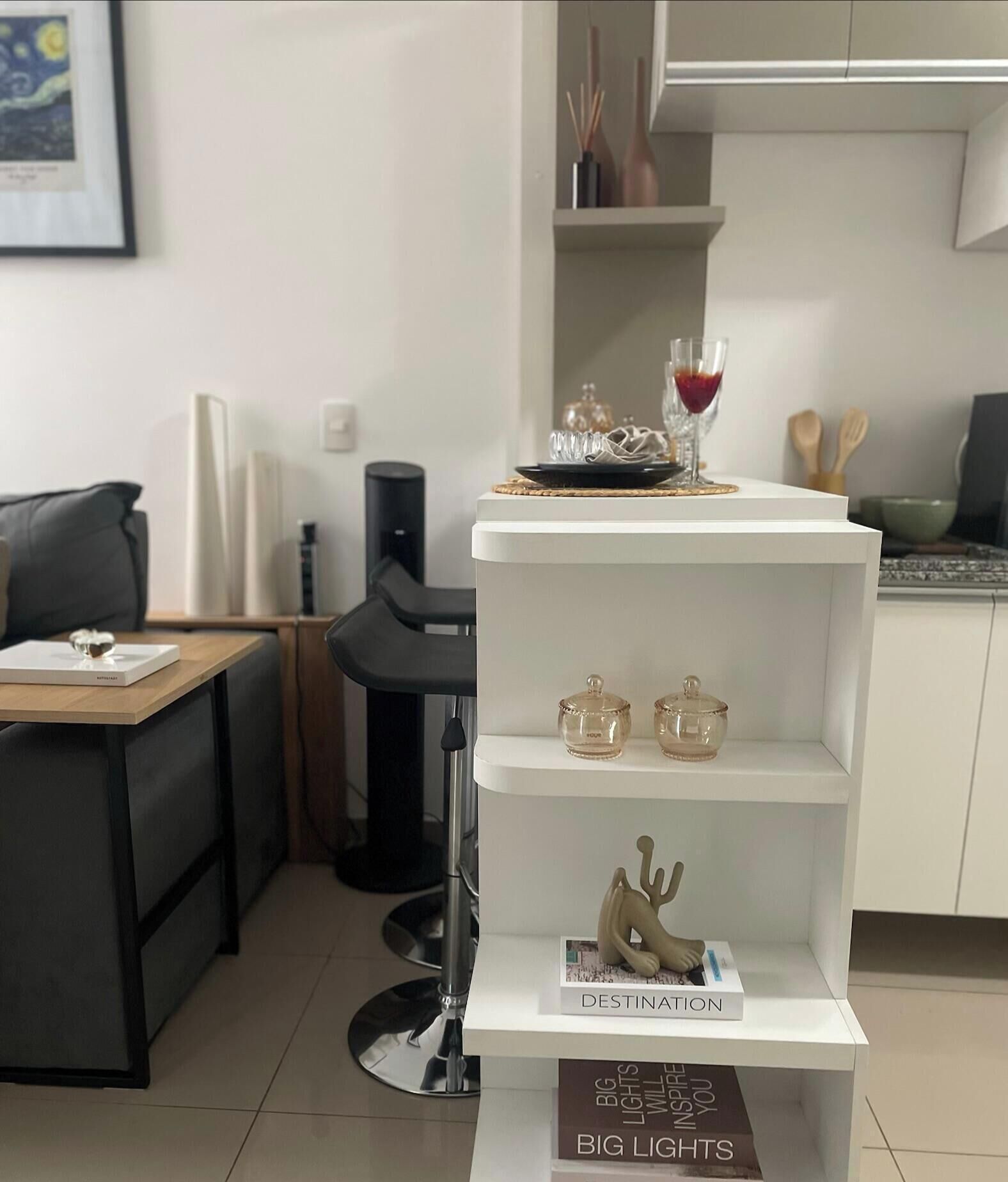 Cozinha moderna com armários brancos, área de jantar com mesa de madeira e cadeiras pretas, e estante branca com objetos decorativos e uma taça de vinho.