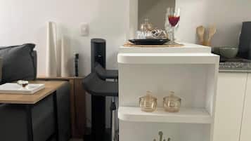 Cozinha moderna com armários brancos, área de jantar com mesa de madeira e cadeiras pretas, e estante branca com objetos decorativos e uma taça de vinho.