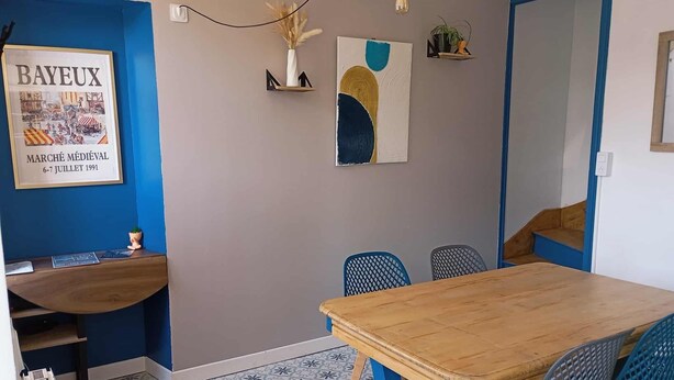 Un comedor con una mesa de madera y sillas azules, un cartel enmarcado en la pared y un pequeño estante con plantas.