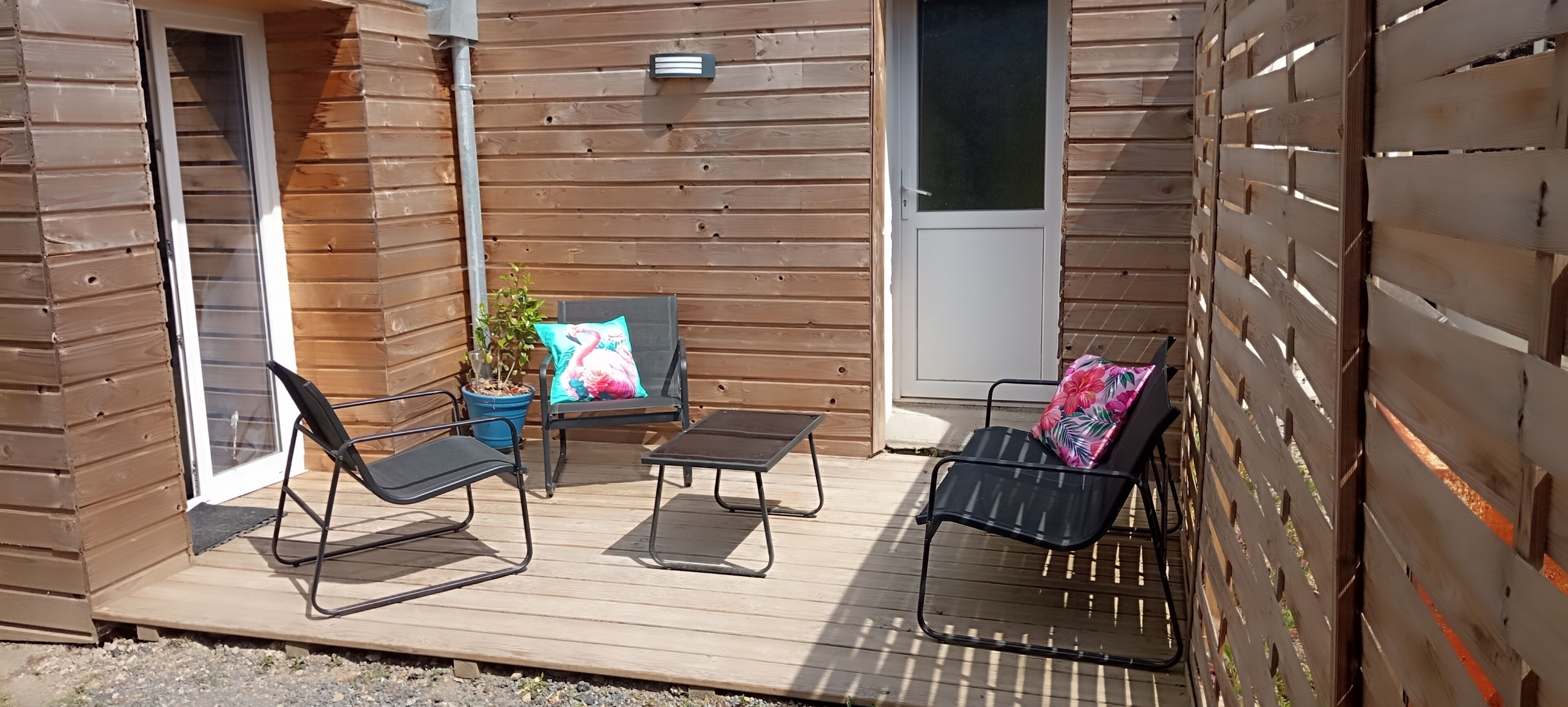 Een houten terras met twee loungestoelen, een bijzettafeltje, een bloempot en een deur.