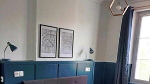 Un dormitorio con cama, cabecera, cómoda, dos lámparas de pared y dos cuadros enmarcados.