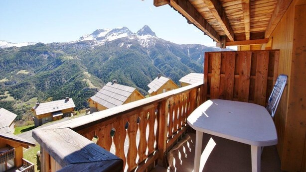 Un balcon en bois, agrémenté d’une table et de chaises, qui offre une vue sur un paysage montagneux parsemé de plusieurs maisons.