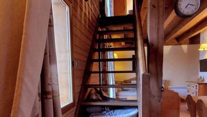 Un escalier en bois mène à une chambre mansardée équipée d’un lit, d’une horloge et d’une fenêtre avec des rideaux.
