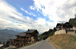Une route de montagne bordée de bâtiments en bois des deux côtés, avec des balcons et une vue sur les montagnes en arrière-plan.