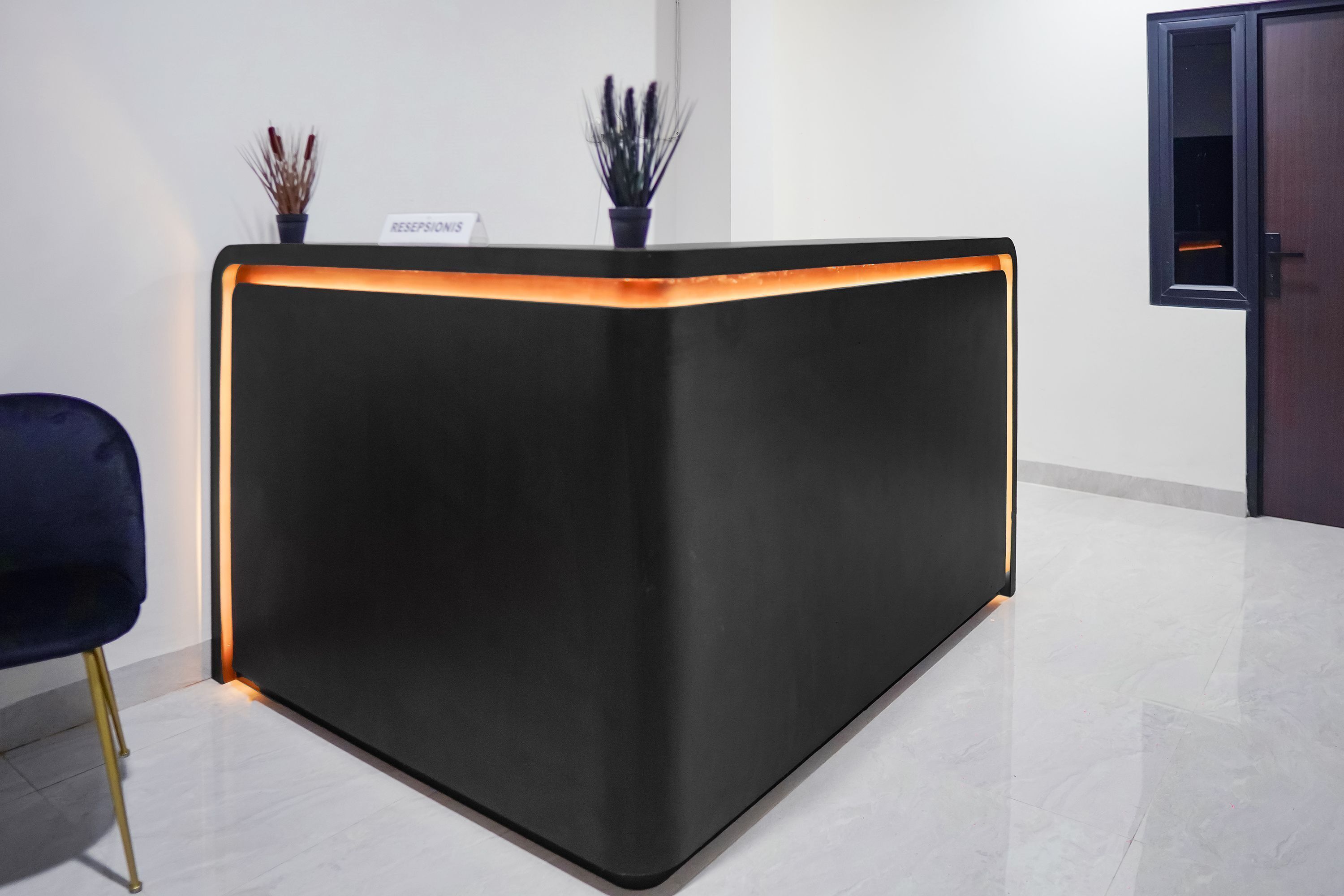 A reception desk with a black finish and a glowing orange light strip. There are two potted plants on top of the desk.