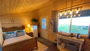 Een houten cabinestijl interieur met een bed, een bureau met een lamp, een raam met uitzicht op de omgeving en een koelkast.