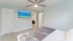 Un dormitorio con ventilador de techo, televisor grande en la pared y vista al océano a través de una puerta abierta.