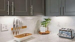 Une cuisine avec des meubles gris, un pare-feu en carreaux blancs, une planche à découper en bois, un saladier blanc, un grille-pain et une plante en pot.