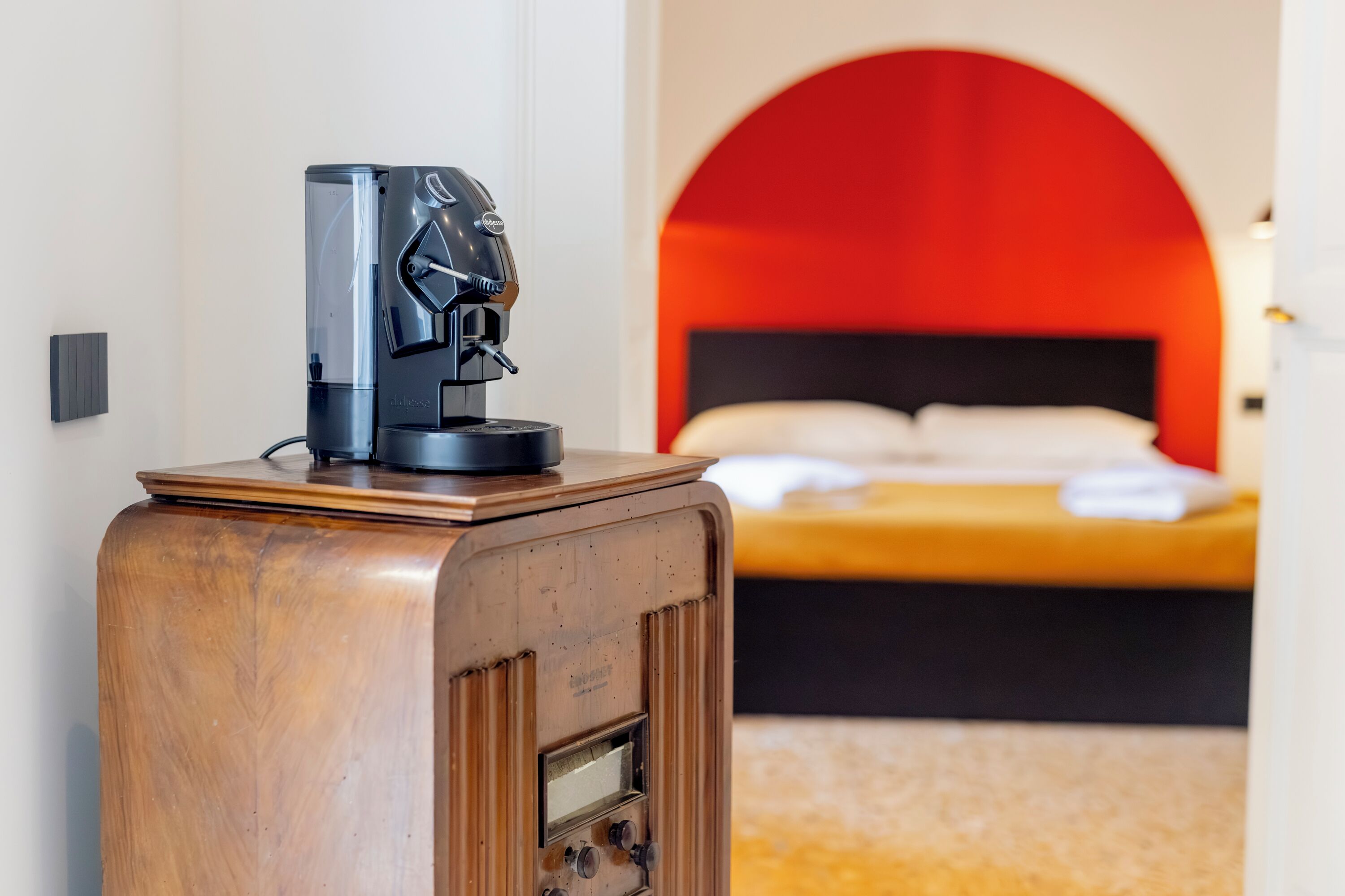 A coffee machine on a wooden cabinet in a hotel room with a bed and a red arch in the background.