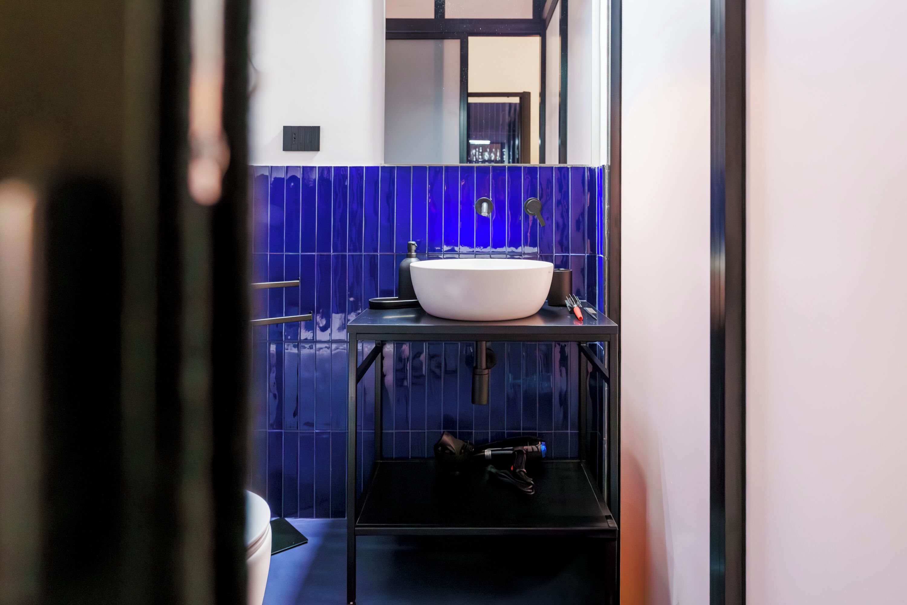 Un baño con un lavamanos blanco sobre un estante negro, paredes con azulejos azules y un espejo sobre el lavamanos.