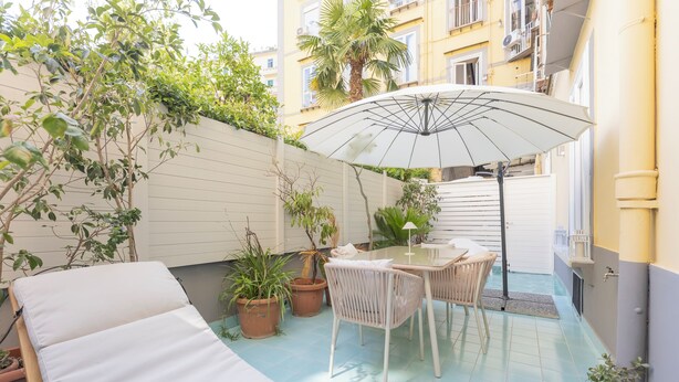 Une terrasse ensoleillée avec un fauteuil de relaxation, une table avec des chaises et des plantes en pot.
